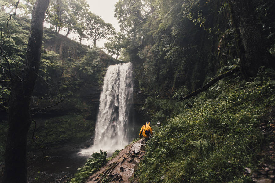 Person Walking Beside Waterfalls Surrounded By Trees Wallpaper
