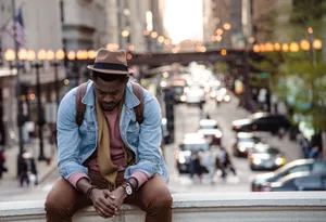 A Man In A Leather Jacket Looking Down While Sitting On A Ledge In A City Wallpaper