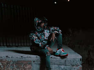 A Man Sitting On A Wall At Night Wearing A Hoodie And Sneakers Wallpaper