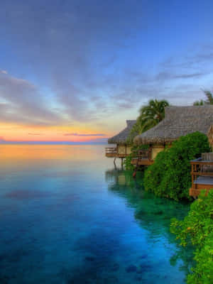 A Tropical Island With Thatched Huts And A Sunset Wallpaper